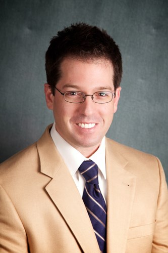 Professional business headshot of a main with dark hair and wire glasses smiling. He is dressed in a tan sportcoat, white shirt, and blue tie.