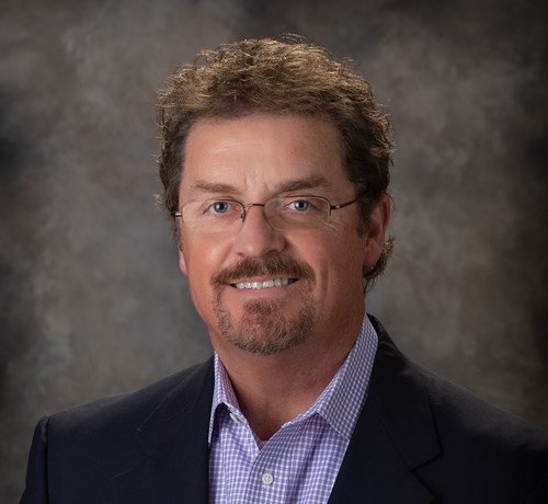 Professional headshot of a caucasian male with light brown hair, a goatee, and eye glasses. He is wearing a blue sportcoat and checkered blue collared shirt.