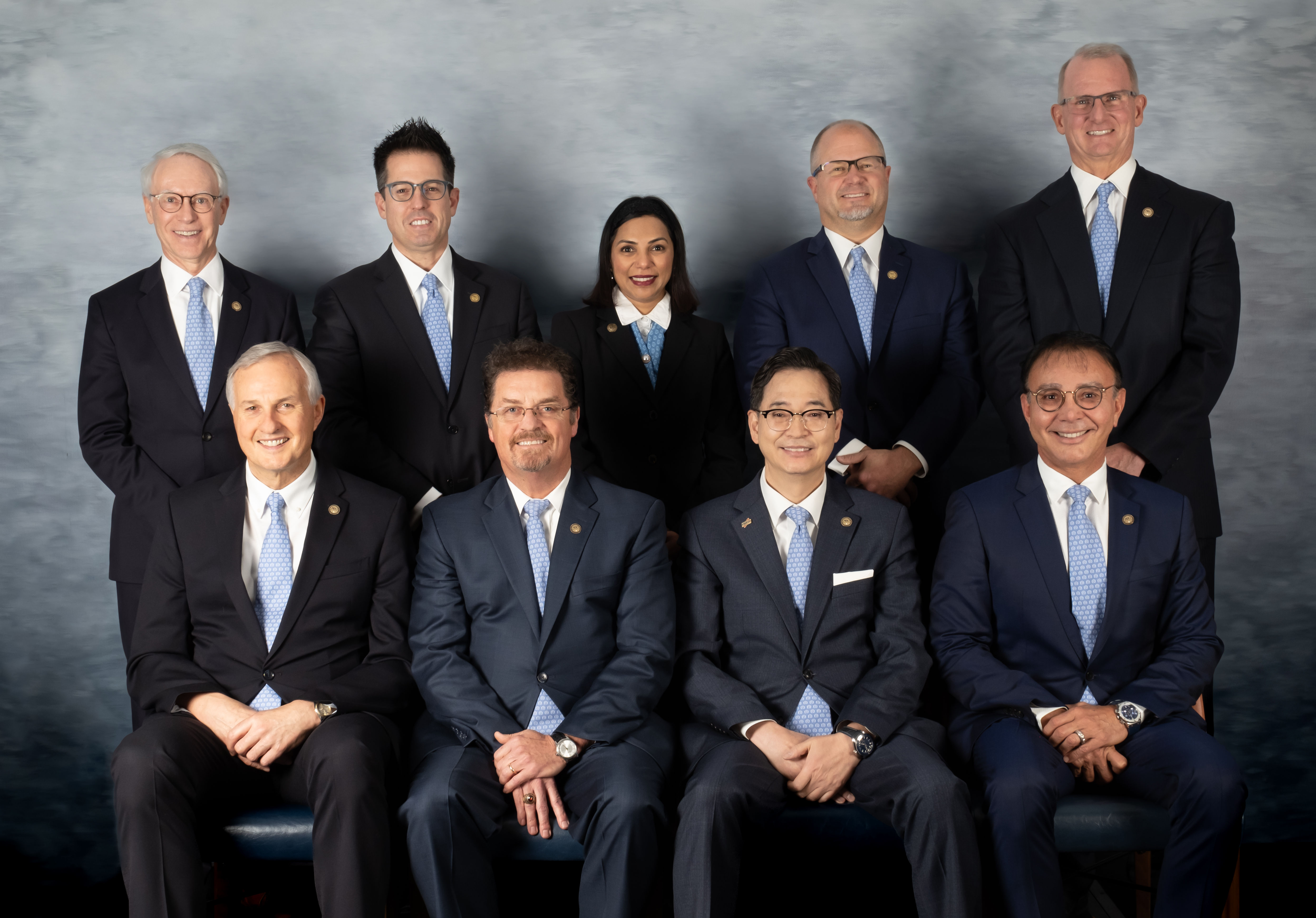 well-lit photo of 8 people wearing blue suits. There are 5 standing in a row behind 4 sitting in chairs.